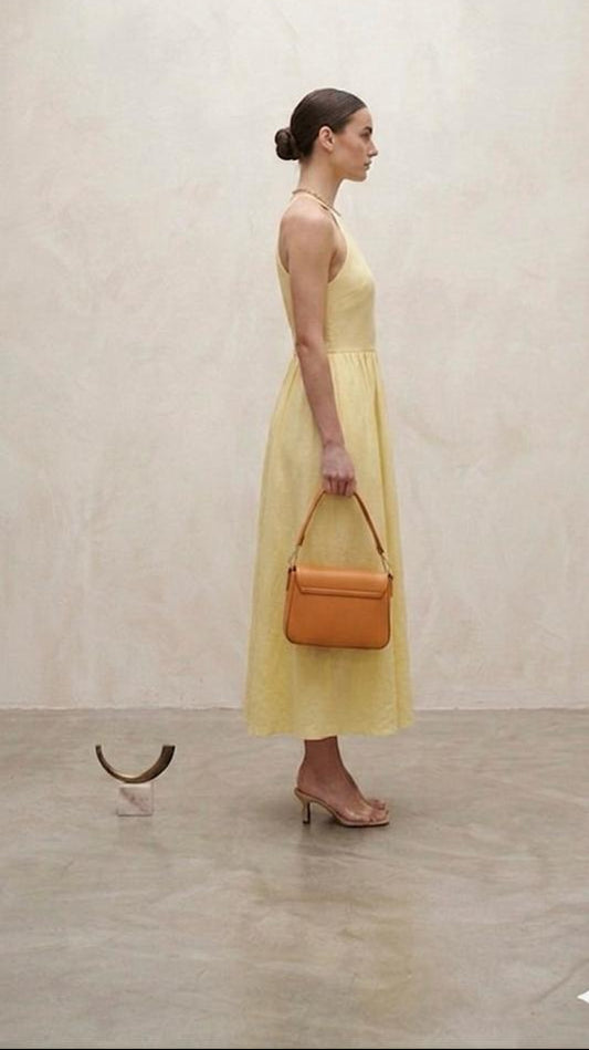 Side view of a pastel yellow halter-neck midi dress paired with nude heels and a tan handbag.”