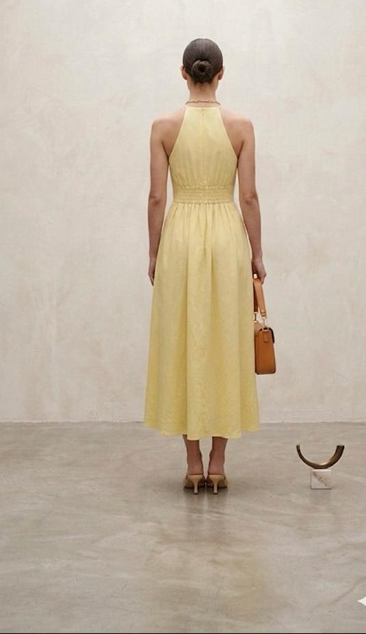 Back view of a pastel yellow halter-neck midi dress with flowing skirt and open shoulder detailing.