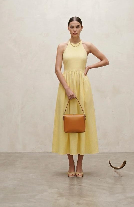 Woman wearing a pastel yellow halter-neck midi dress with nude heels and a tan leather handbag, front view.